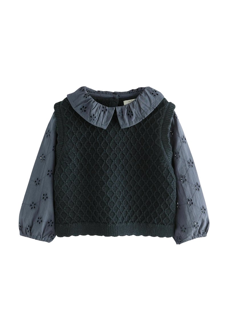 Pull sans manches tricoté vert foncé porté sur une blouse grise à manches longues avec des motifs floraux découpés et un col à volants.