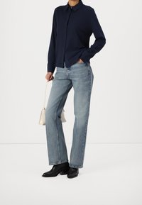 Granatowa koszula z długim rękawem zapinana na guziki w połączeniu z jasnoniebieskimi dżinsami o prostym kroju i czarnymi botkami na obcasie. Modelka trzyma małą beżową torbę.