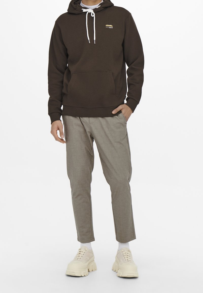 Hombre vestido con sudadera marrón, pantalones de cuadros grises, calcetines blancos y zapatillas beige robustas, de pie con una mano en el bolsillo sobre un fondo blanco.