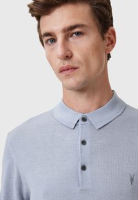 Ljusblå polo skjorta av mjukt tyg, med klassisk krage, tre mörka knappar och en liten logotyp på vänster bröst.