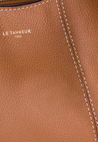 Braune Lederstruktur mit feiner Narbung, hervorgehoben durch weiße Steppnähte entlang einer Naht. Das Markenlogo "LE TANNEUR 1836" ist geprägt.