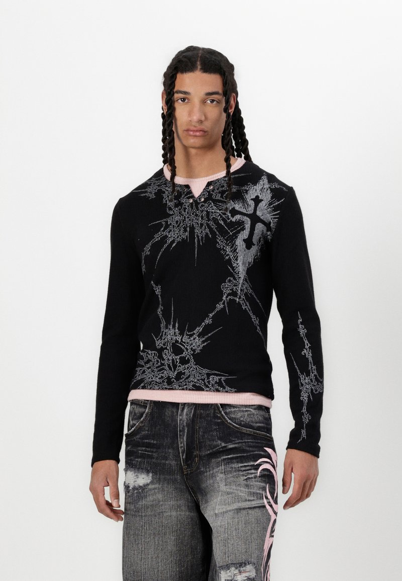 Schwarzer gerippter Pullover mit langen Ärmeln und grafischem silbernem Design, kontrastierender pinker Kragen, kombiniert mit destroyed grauen Jeans mit pinken Akzenten.
