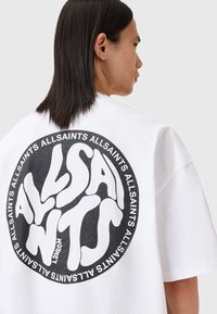 Osoba s dugom tamnom kosom okrenutom bočnoj strani, koja nosi bijelu majicu s velikim crnim kružnim logom "AllSaints London" na leđima.