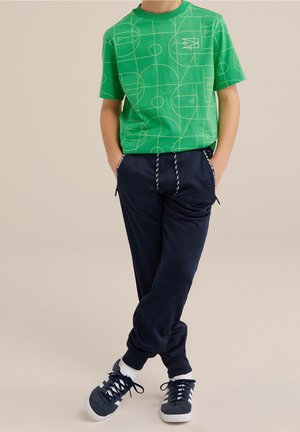 Child wearing green t-shirt with geometric pattern, navy jogger pants with drawstring, white socks, and black and white sneakers, standing with hands in pockets.