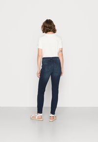 Jeans de mezclilla azul oscuro, ajustados y de cintura alta, con bolsillos traseros y costuras sutiles, combinados con una camiseta blanca básica y sandalias.