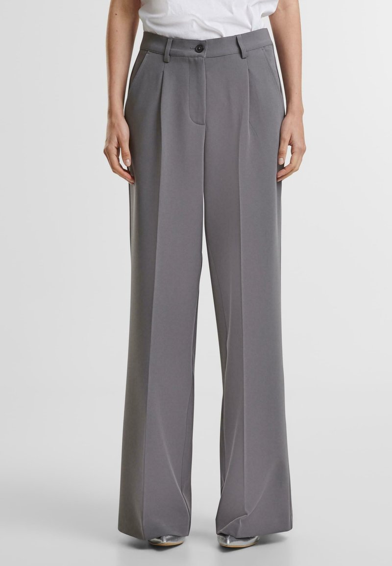 Personne portant un pantalon gris taille haute à jambes larges avec plis sur le devant et fermeture par bouton, associé à un haut blanc et des chaussures argentées.