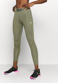 Legginsy oliwkowe z wysokim stanem z logo "PUMA" w kolorze żółtym. Gładka tekstura, dopasowany krój, zestawione z czarnymi butami z różowymi akcentami.