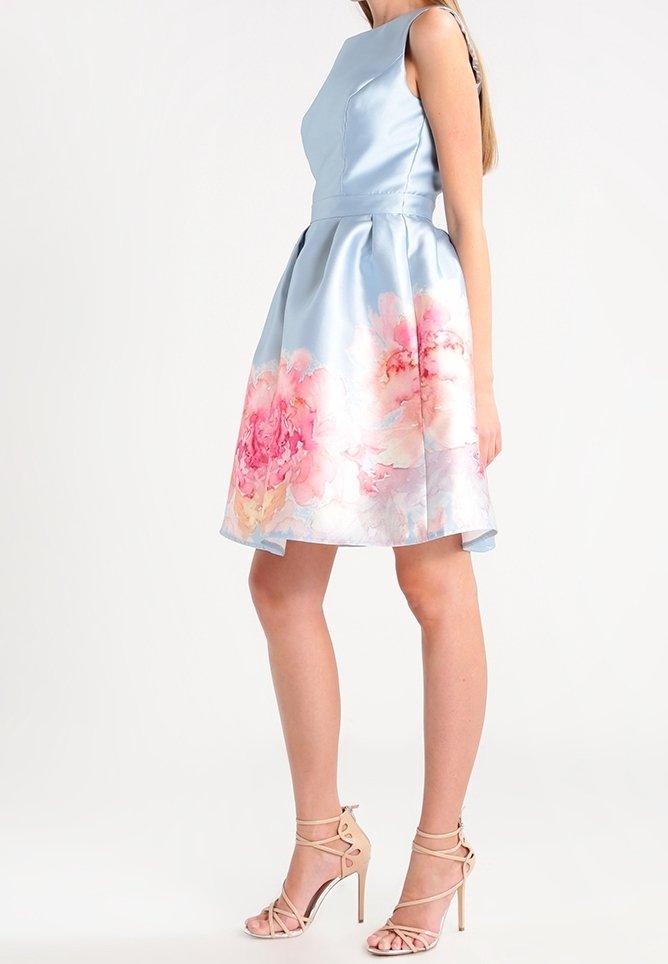 Robe en satin bleu clair avec un corsage ajusté et une jupe évasée, ornée de motifs floraux roses au niveau de l'ourlet. Associée à des escarpins nus à lanières.