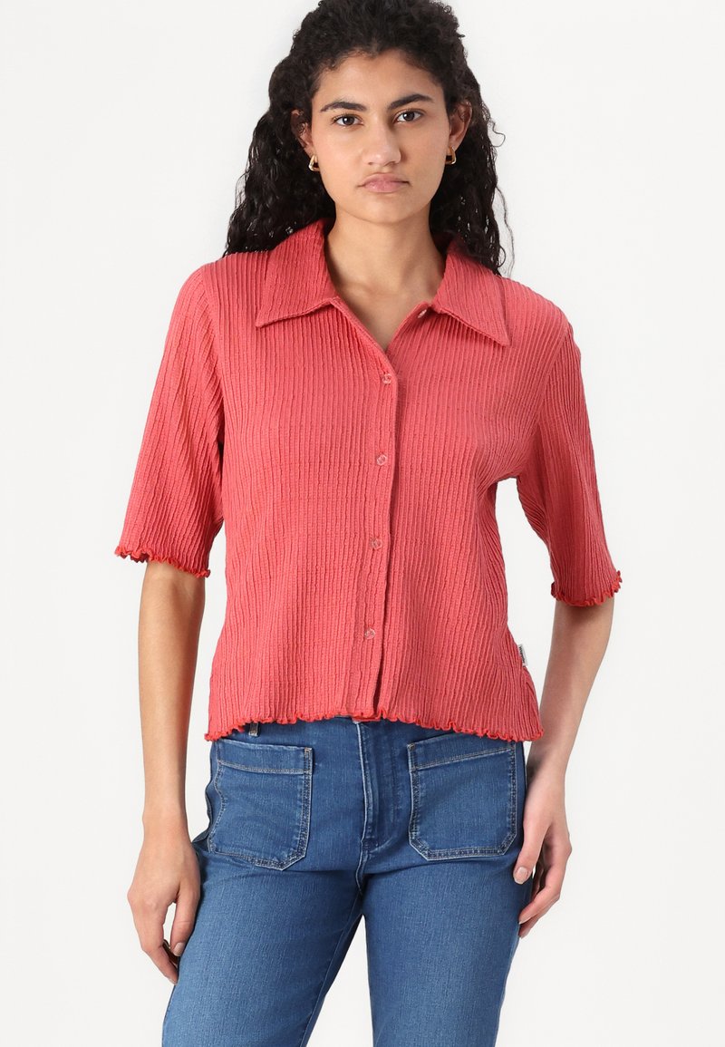 Camicetta corta a maniche corte di colore corallo con bottoni davanti, colletto a punta e bordi scallop. Abbinata a jeans in denim blu classici.