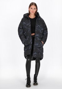 Padded schwarze Jacke mit blauem Marmor-Muster, Kapuze und Fronttaschen. Getragen über einem kurzen Oberteil und schwarzen Leggings, kombiniert mit Schnürstiefeln.