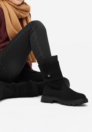 Persona che indossa stivaletti neri testurizzati con dettaglio di bottoni, jeans neri, maglione color bordeaux e sciarpa color cammello, seduta su una superficie bianca.