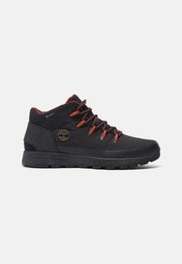 Chaussure de randonnée noire mi-montante avec lacets rouges, semelle texturée et petit logo arbre doré sur le côté.