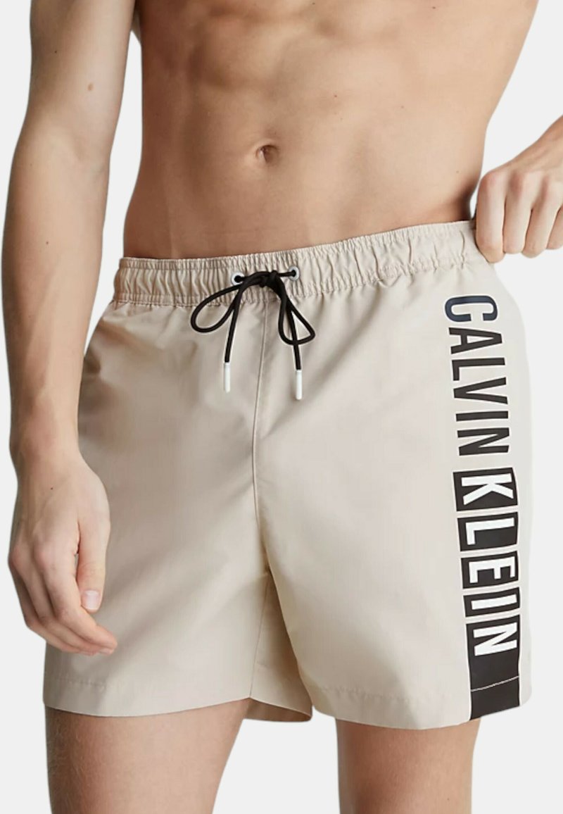 Pantaloni da bagno beige con coulisse nera e logo "CALVIN KLEIN" stampato di lato. Tessuto liscio, lunghezza a metà coscia e vita elasticizzata.