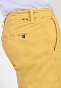 Pantalons en coton jaune au toucher lisse, comportant une poche arrière et un détail de marque. Des passants de ceinture et un léger éclat sont visibles.