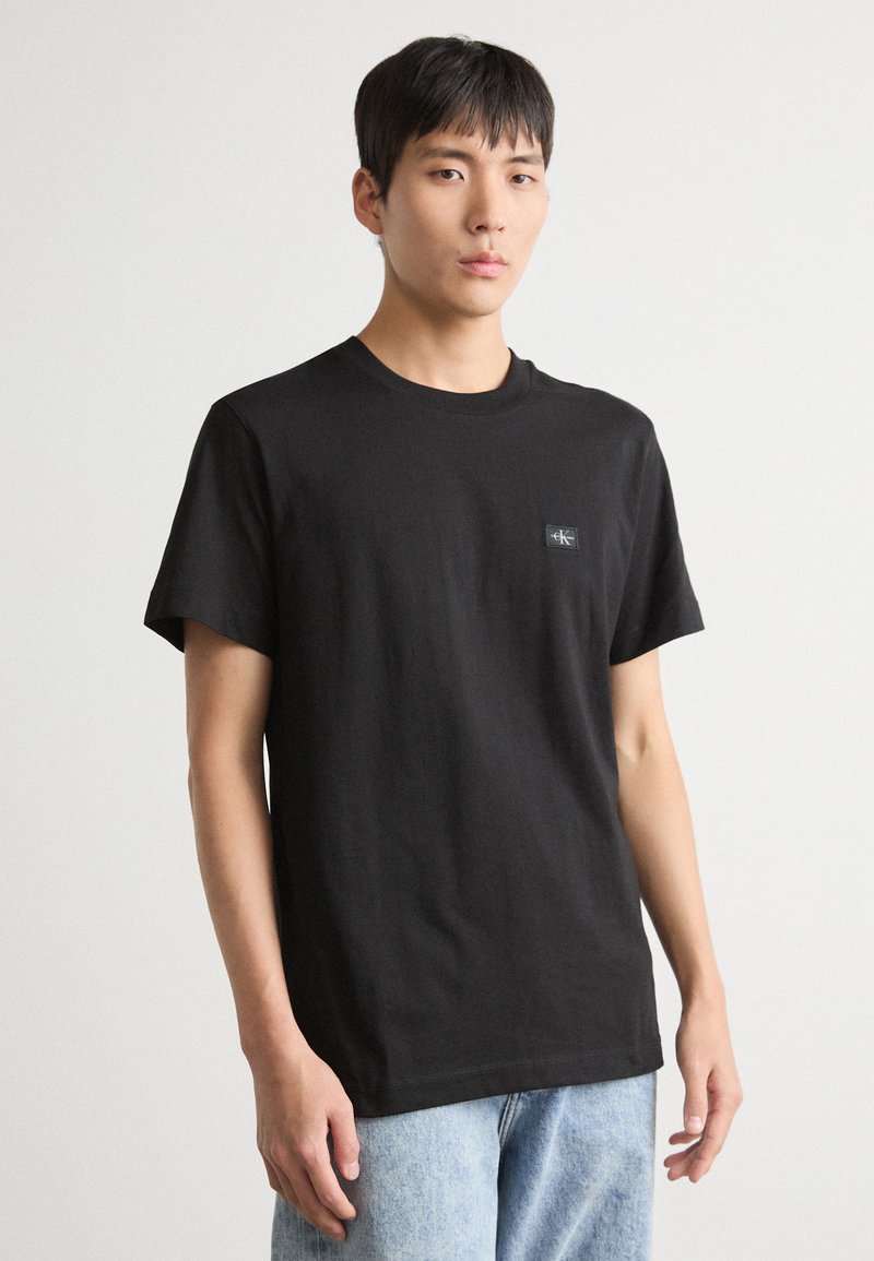 Black cotton t-shirt with a crew neck and short sleeves, featuring a small logo patch on the left chest. Worn with light blue jeans.