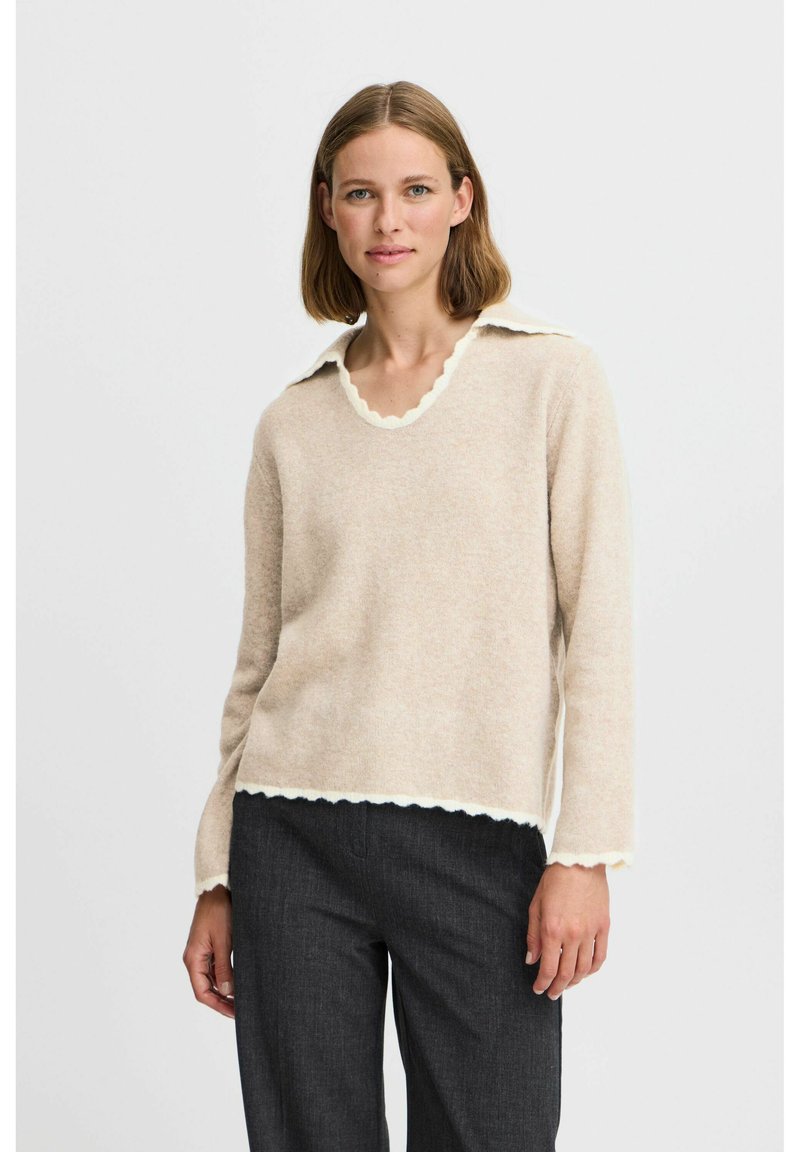 Beige tröja i mjukt material med en scallopad vit krage och nederkant, med långa ärmar och en avslappnad passform. Matchas med mörka byxor.
