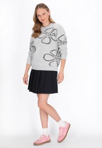 Junge Frau in einem grauen Pullover mit schwarzen Blumenmustern, einem schwarzen Faltenrock, weißen Socken und pinken Sneakern, die vor einem weißen Hintergrund steht.