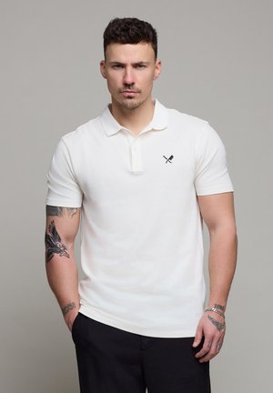 Mann med kort mørkt hår og tatoveringer, iført hvit polo-skjorte med logo av kryssende balltre og ball, står mot en grå bakgrunn.