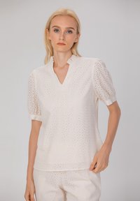 Blouse blanche à manches courtes avec un col en V, un motif de perforation et des accents à volants au col. Le tissu semble léger et respirant.