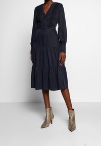 Kvinna klädd i en marinblå långärmad midiklänning med spetsdetaljer och volangkjol, i kombination med spetsiga ankelboots i ormskinn.