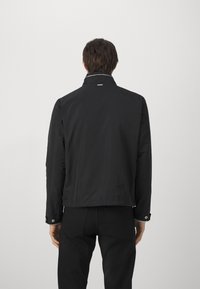 Svart lett jakke med ståkrage, justerbare mansjetter og glatt tekstur. Enkelt design, ingen synlige mønstre eller aksenter.