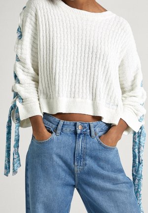 Witte ribgebreide trui met blauwe bloemenstrikjes aan de mouwen, gecombineerd met lichtblauwe high-waisted jeans. De trui heeft een cropped ontwerp en brede manchetten.