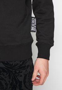 Svart bomullströja med ribbstickade ärmslut och nederkant. Har en framträdande vit etikett med texten "VERSACE JEANS COUTURE" på sidan.