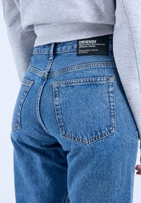 Blue denim jeans with a classic fit, featuring a black tag at the waist. Five pockets and visible stitching details on a textured fabric.