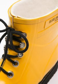 Bota impermeable amarilla con cordones negros, ojales de metal y forro de lana blanca, mostrando detalles laterales y superiores.