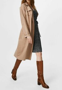 Jasnobrązowy zamszowy płaszcz trench z długimi rękawami, nałożony na ciemną sukienkę w małe kwiatowe wzory, zestawiony z brązowymi kozakami do kolan.