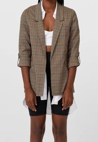 Hnědý kostkovaný blejzr s uvolněným střihem, ohrnutými rukávy a strukturovanými klopami, přes bílé zkrácené tričko a černé šortky. Textura z bavlněné směsi.