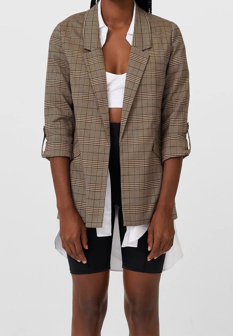Hnědý kostkovaný blejzr s uvolněným střihem, ohrnutými rukávy a strukturovanými klopami, přes bílé zkrácené tričko a černé šortky. Textura z bavlněné směsi.
