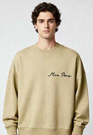 Jeune homme aux cheveux ondulés et foncés portant un sweat-shirt beige brodé de "Mira Paris" en écriture noire.