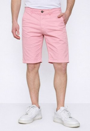 Homme portant un short rose clair jusqu'aux genoux et des baskets blanches, debout avec une main dans la poche devant un fond uni.