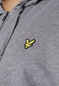 Gros plan d'une personne portant un sweat à capuche gris zippé avec un petit emblème d'oiseau jaune sur la poitrine.
