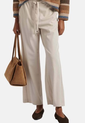 Persoon draagt beige broek met wijde pijpen, bruine platte schoenen, een bruin gestreepte trui en houdt een bruine suède handtas naast zich vast.