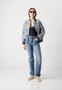 Bloemmotief bomberjack met marineblauwe accenten, zwarte top, lichtblauwe jeans en zilveren sneakers. Model draagt donkere zonnebril.
