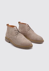Botas de tobillo de ante beige con punteras redondas, cordones en la parte delantera y costuras amarillas contrastantes a lo largo de la suela. Forro interior de cuero suave en color marrón claro.