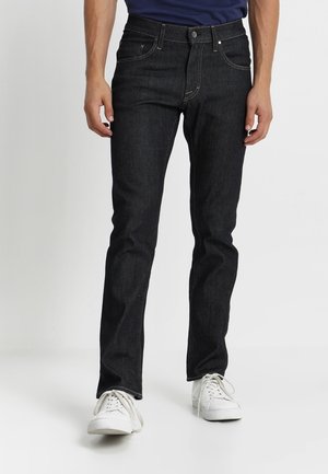 Homme portant un jean slim foncé et des baskets blanches, debout devant un fond blanc uni.