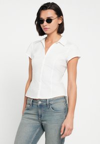 Άσπρο πουκάμισο με κοντά μανίκια και κουμπιά, εφαρμοστό σχέδιο με μυτερό γιακά, συνδυασμένο με ανοιχτό μπλε τζιν. Λεία υφή, απλή κατασκευή.