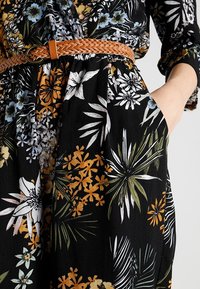 Jumpsuit floreale in tessuto nero, con fiori bianchi, gialli e blu, dotato di una cintura intrecciata marrone in vita e tasche laterali.