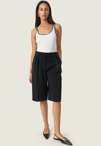 Schwarze, taillierte Shorts mit Falten, kombiniert mit einem weißen Tanktop mit schwarzem Saum und schwarzen, spitzen Ballerinas. Einfaches und strukturiertes Design.