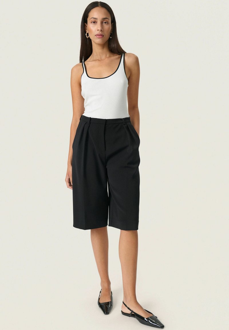 Pantalones cortos negros ajustados con pliegues, combinados con una camiseta sin mangas blanca con ribete negro y zapatos planos negros con punta. Diseño simple y estructurado.