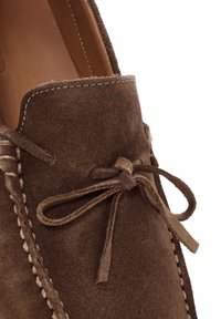 Zapato mocasín de ante marrón con un acento cosido, punta redonda y forro de cuero. Presenta un detalle de lazo y una superficie texturizada.