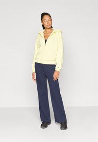 Ανοιχτό κίτρινο φούτερ με φερμουάρ, κουκούλα και μπροστινές τσέπες, συνδυασμένο με σκούρο μπλε παντελόνι φαρδιάς γραμμής και μαύρα παπούτσια.