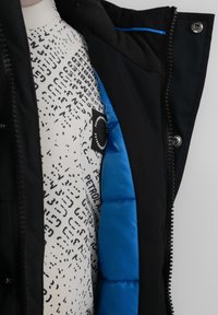 Svart ytterjacka med ett vadderat foder, med en blå insida. Vit innertröja med svart abstrakt tryck och logotypdetalj.