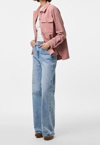 Persoon in een roze corduroy jasje over een witte top, lichte blauwe hoge wijde jeans en bruine schoenen, staand met de handen in de zakken.
