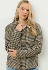 Giacca a motivo houndstooth marrone e beige con colletto zip, maniche lunghe e tasche laterali, realizzata in un materiale tessuto e strutturato.