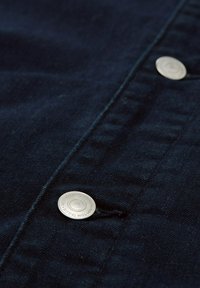 Marineblauer Stoff mit glatter Textur, versehen mit silbernen Metallknöpfen. Sichtbare Nahtdetails entlang des Randes und der Knopfleiste.