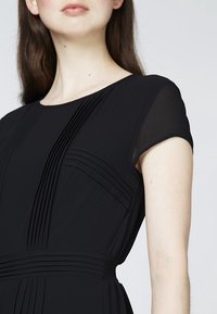 Schwarzes Kleid mit vertikalen Falten, kurzen durchsichtigen Ärmeln und einem gerafften Taillenbereich. Glattes Stoff mit strukturierter Form und klassischem Rundhalsausschnitt.